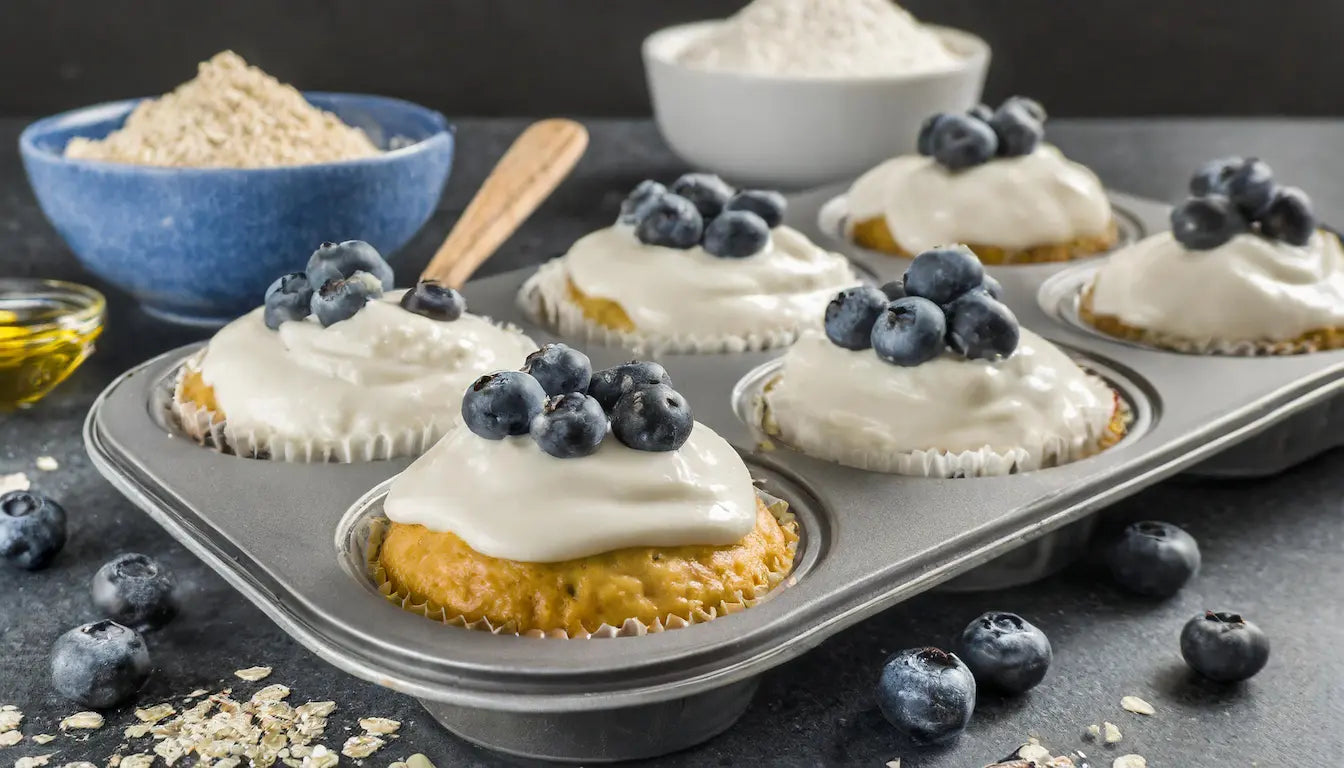 Muffins de yogur cubiertos con arándanos frescos, servidos en una bandeja para hornear, con avena, harina y miel como ingredientes visibles en el fondo.