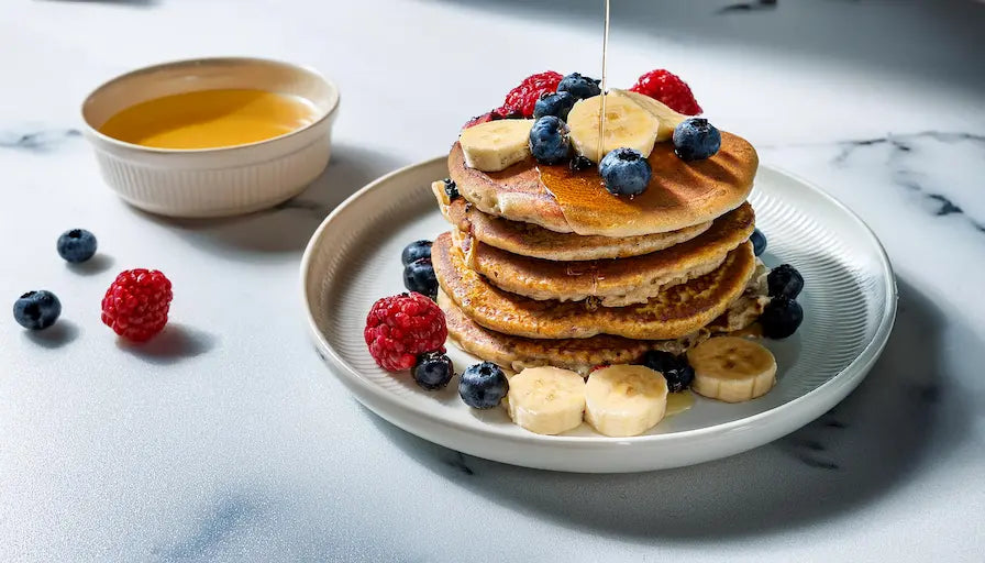 Torre de panqueques de calabaza y especias cubiertos con nueces y almendras, acompañados de jarra de leche, miel de calabaza, canela y una calabaza en el fondo.