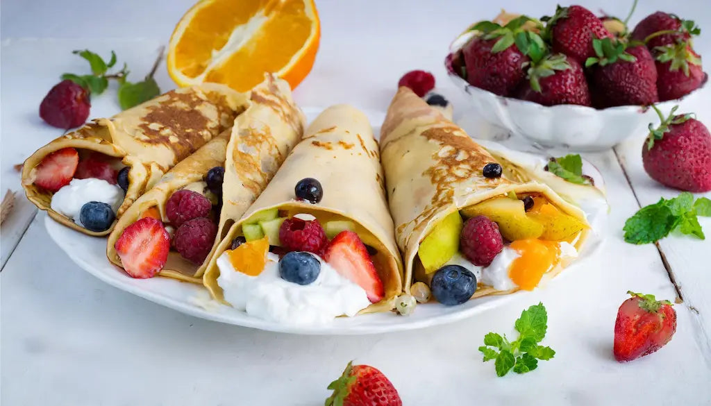 Crepas de proteína rellenas de frutas frescas como fresas, arándanos, frambuesas y rodajas de naranja, servidas en un plato blanco con una guarnición de fresas y menta.